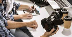 Alunos praticando edição de imagens e manuseio de câmeras profissionais durante curso presencial de fotografia, com laptops e equipamentos fotográficos sobre a mesa.