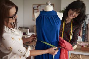 Alunas durante aula prática de um curso presencial de modelagem, tirando medidas em um manequim com vestido azul em um ateliê de costura.