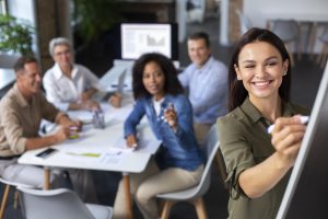 Grupo de adultos participando de uma aula prática durante cursos presenciais, com uma mulher sorridente escrevendo em um quadro e colegas atentos em uma sala de treinamento.