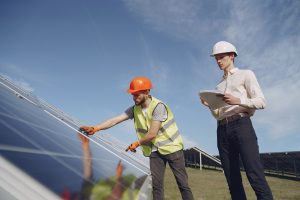 Dois técnicos em campo realizando a instalação e inspeção de painéis solares em uma usina de energia solar, usando equipamentos de segurança.