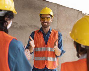 Instrutor de segurança do trabalho orientando equipe em obra, todos usando capacete, colete refletivo e equipamentos de proteção individual.